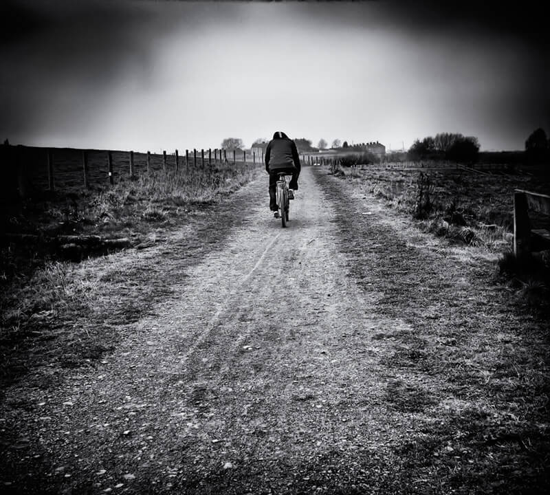 A monochrome capture of a cyclist pedaling down an unpaved path.