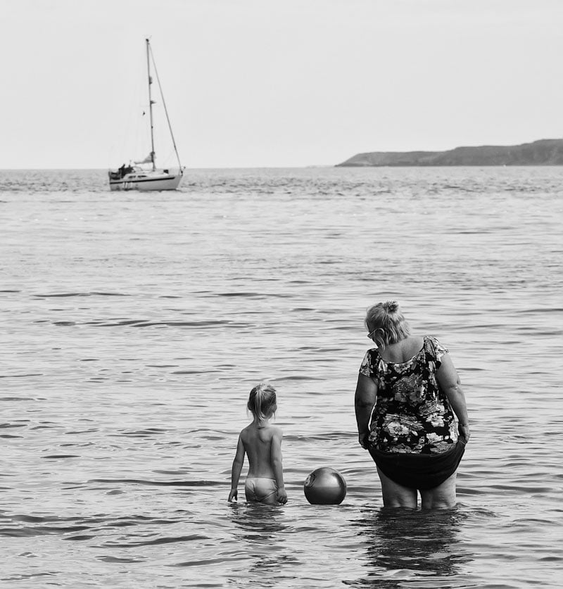 A woman and child enjoying The Coast in the water with a sailboat in the background.