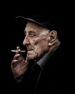 A street photography capturing an old man smoking a cigarette against a black background.