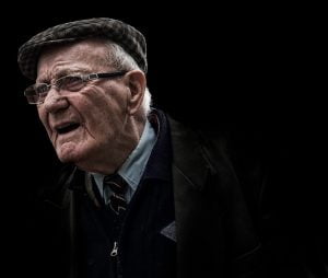 A street photographer captures an elderly man in a hat and jacket against a black backdrop.