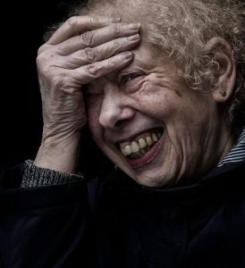 Street photography of an old woman laughing with her hands on her face.
