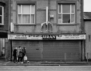 A black and white street photography capturing people outside a pizza shop.