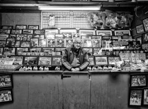 A black and white street photography capturing a man standing behind a counter full of framed pictures.