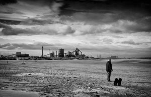 A man captured in a street photography style, standing on a beach with a dog in the background.