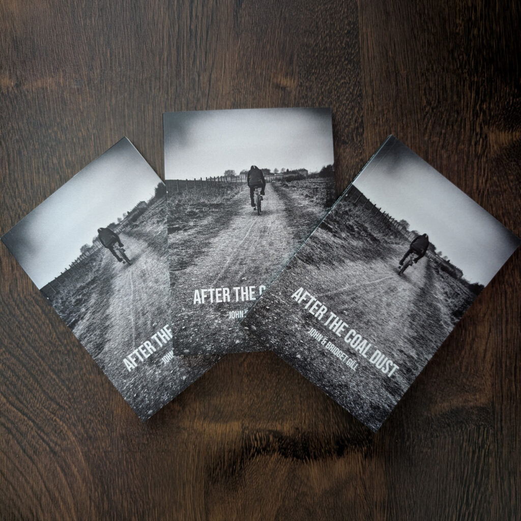 Three copies of the new edition of “After the Coal Dust Book,” each featuring a striking black-and-white cover image of a person walking down a dirt path, are displayed on a dark wooden surface. This A5 edition brings fresh appeal to the acclaimed work.