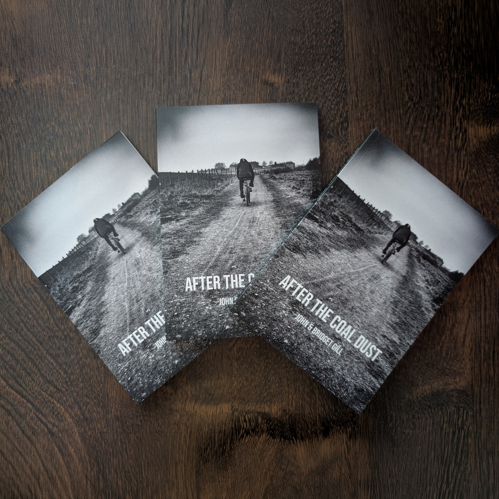 Three copies of the new edition of “After the Coal Dust Book,” each featuring a striking black-and-white cover image of a person walking down a dirt path, are displayed on a dark wooden surface. This A5 edition brings fresh appeal to the acclaimed work.