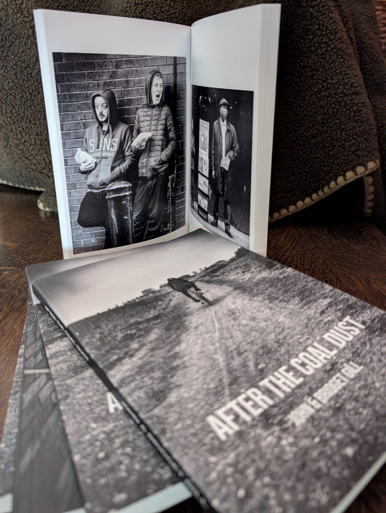 Three copies of the A5 edition of “After the Coal Dust Book” are displayed, with one open to a page showing a black-and-white image of two men standing against a brick wall.